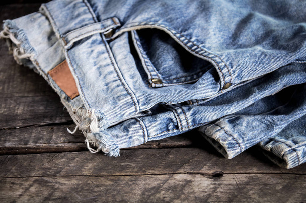 Jeans gestapeld op een houten achtergrond. Blauwe jeans op een bruine houten achtergrond. Gerafeld jeans. Detail van mooie blauwe jeans met lederen riem in vintage stijl. Textuur van blauwe jeans achtergrond - Foto, afbeelding