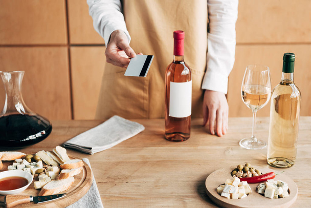 vista ritagliata del sommelier in grembiule in piedi vicino al tavolo con bottiglie di vino e in possesso di carta di credito
 - Foto, immagini