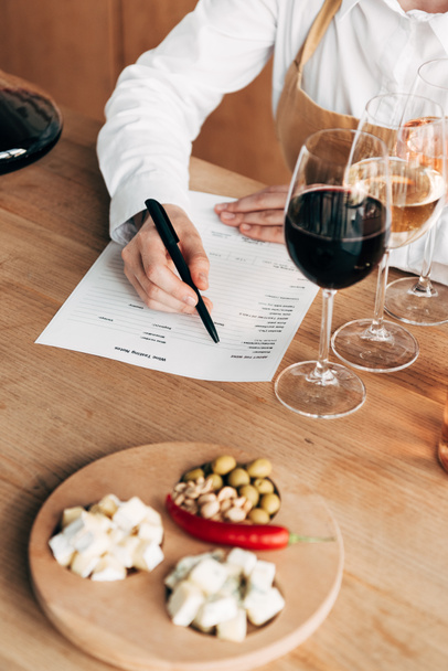 обрезанный вид сомелье в фартуке сидит за столом и писать в вино дегустации документа
 - Фото, изображение