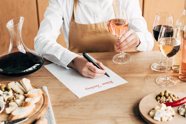 cropped view of sommelier in apron sitting at table, holding wine glass and writing in wine tasting document - Fotó, kép