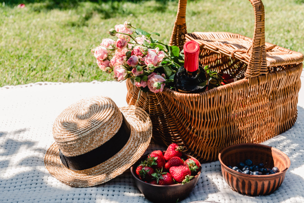 cesta de vime com rosas e garrafa de vinho em cobertor branco perto de chapéu de palha e bagas no dia ensolarado no jardim
 - Foto, Imagem