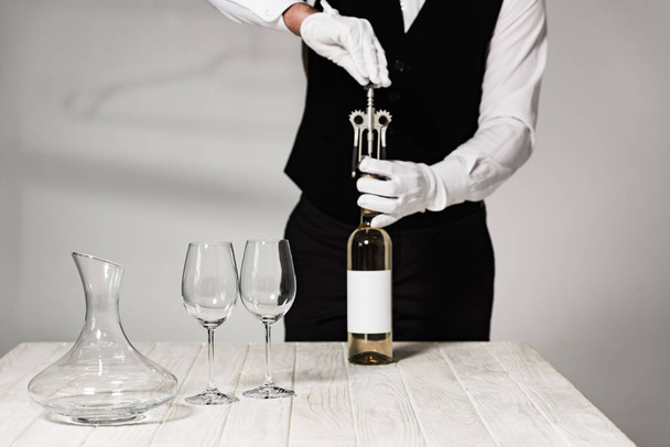 částečný pohled na číšníka v bílých rukavicích otevírající láhev vína s korkšroubem - Fotografie, Obrázek