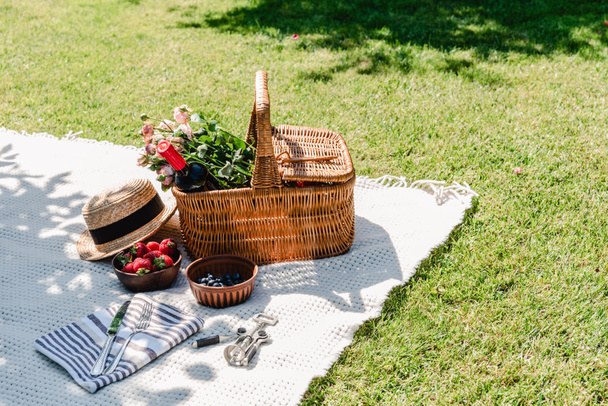 wicker basket with roses and bottle of wine on white blanket near straw hat, cutlery on napkin and berries at sunny day in garden - Photo, Image