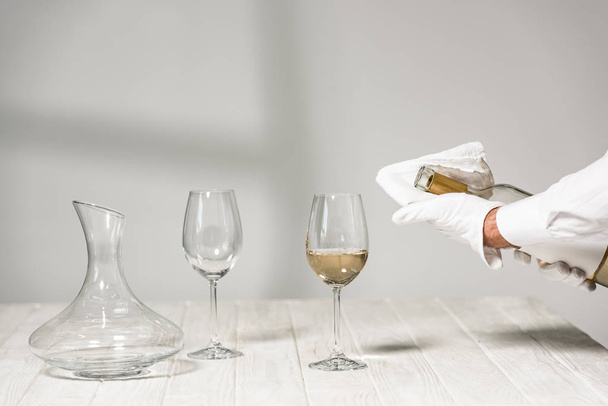 cropped view of water in white glove holding bottle of wine near wine glasses on wooden table - Photo, image