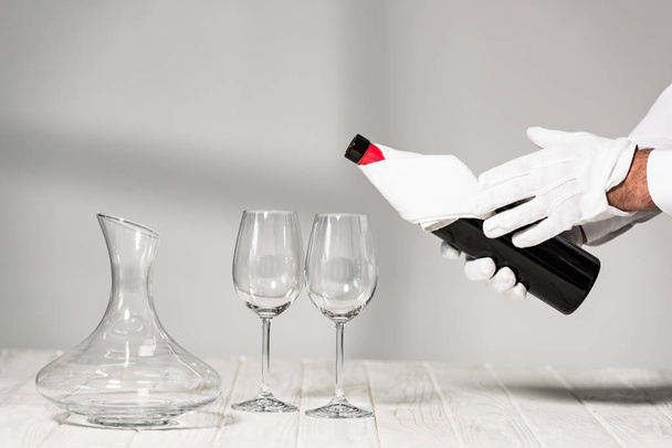 oříznutý pohled na vodu v bílých rukavicích držící láhev vína u skleniček na dřevěném stole - Fotografie, Obrázek