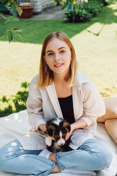 happy blonde girl sitting on blanket in garden with adorable puppy at sunny day - Photo, Image