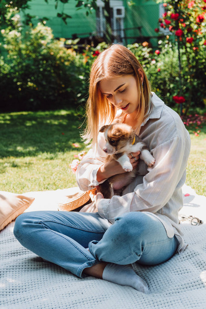 blonde girl sitting on blanket in garden with crossed legs and adorable puppy at sunny day - 写真・画像