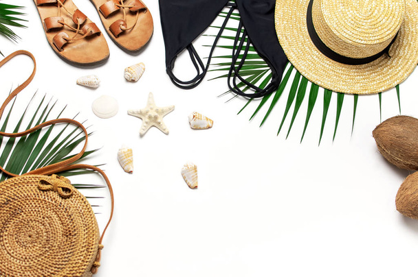 Woman's beach accessories flat lay. Round trendy rattan bag straw hat black swimsuit leather sandals tropical palm leaves coconut seashells on white background. Top view copy space. Summer backdrop - Photo, Image