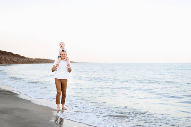 Padre con un figlio biondo sulle spalle cammina lungo la riva del mare. Vacanza in famiglia
 - Foto, immagini