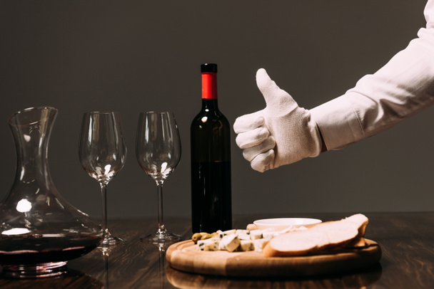 Ausgeschnittene Ansicht des Kellners im weißen Handschuh, der den Daumen in der Nähe des Tisches mit Essen und Wein zeigt - Foto, Bild