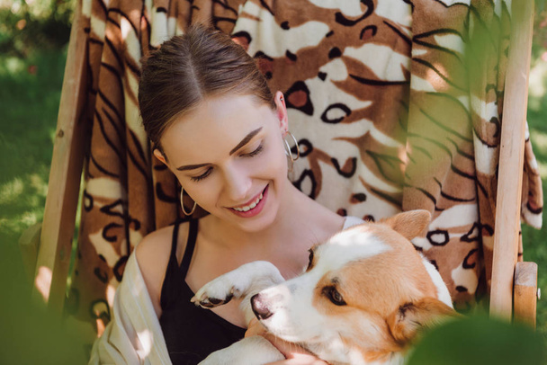 selective focus of green leaves and happy blonde girl holding corgi dog while sitting in deck chair in garden - Фото, изображение