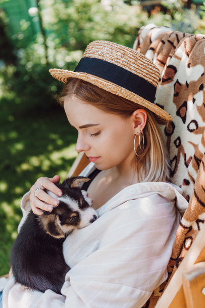 blonde girl in straw hat holding puppy while sitting in deck chair in garden - Photo, Image