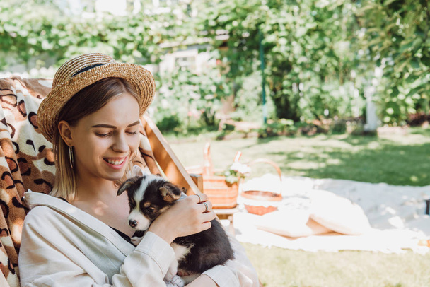 smiling blonde girl in straw hat holding puppy while sitting in deck chair in green garden - 写真・画像
