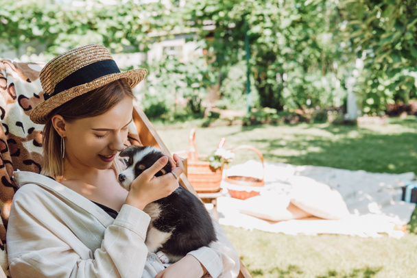 happy blonde girl in straw hat holding puppy while sitting in deck chair in green garden - 写真・画像
