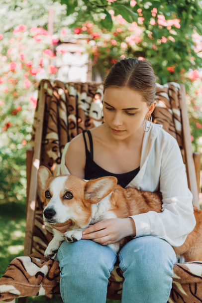 blonde girl holding corgi dog while sitting in deck chair in green garden - Fotografie, Obrázek