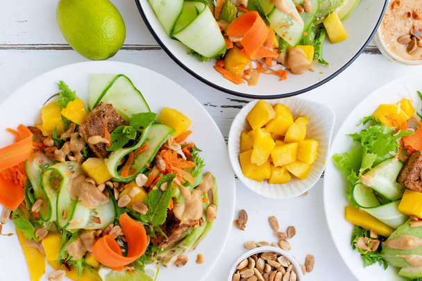 plates with healthy summer fresh salad of mango and avocado with tofu and sweet peanut butter on white wooden table  - 写真・画像
