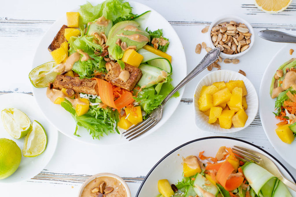 plates with healthy summer fresh salad of mango and avocado with tofu and sweet peanut butter on white wooden table  - Foto, imagen