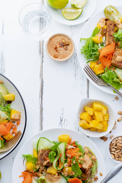 白い木製のテーブルに豆腐と甘いピーナッツバターとマンゴーとアボカドの健康的な夏の新鮮なサラダとプレート  - 写真・画像