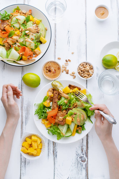 Female hands holding fork over plate of fresh salad with mango and avocado with tofu and sweet peanut butter on white wooden table - Foto, afbeelding