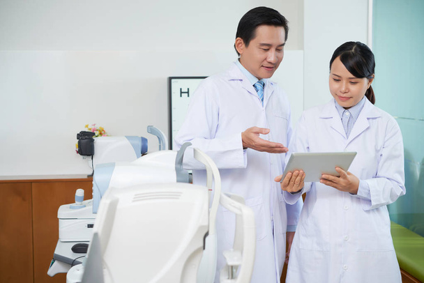 Двоє офтальмологів, чоловік і жінка, стоять у білих халатах і обговорюють використання цифрових планшетів у своїй роботі в офтальмологічній клініці
  - Фото, зображення