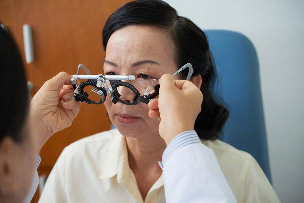 Asian senior woman sitting at optometrist's office while doctor wearing optometrist trial frame on her face to check her eyesight - Photo, Image