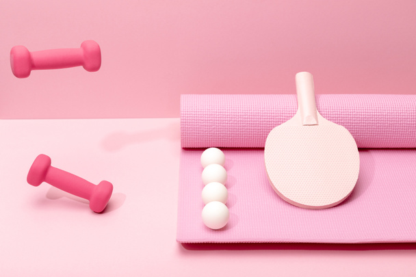pink dumbbells levitating in air near white ping-pong balls and racket on fitness mat on pink background - Photo, Image