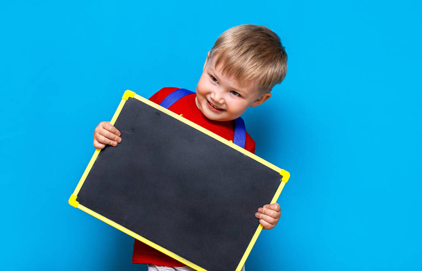 niño pequeño sosteniendo pizarra de tiza de pie sobre fondo azul con bolsa de escuela
 - Foto, imagen