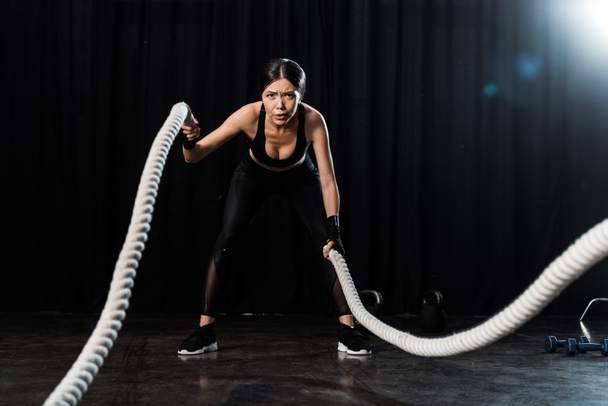 selective focus of attractive sportswoman exercising with battle ropes on black  - Foto, Bild