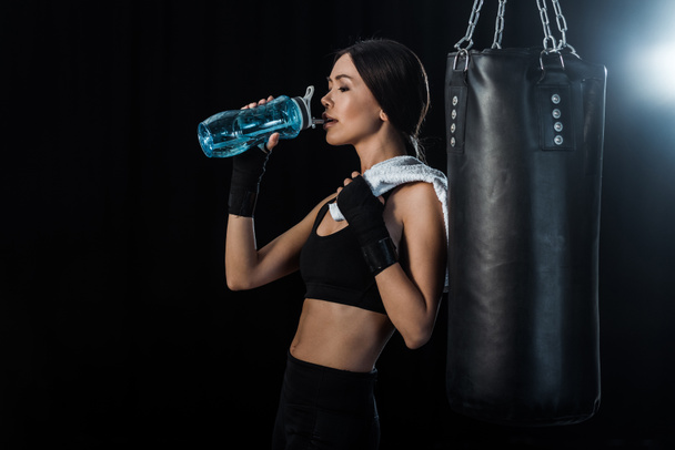 Mädchen trinkt Wasser aus Sportflasche und steht in der Nähe von Boxsack isoliert auf schwarz  - Foto, Bild