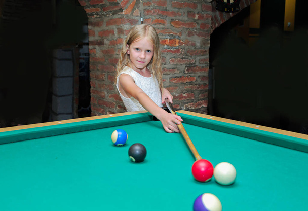 Little beautiful blond girl learns to play billiards, pool, snooker, Russian pyramid in kid's club - Photo, Image