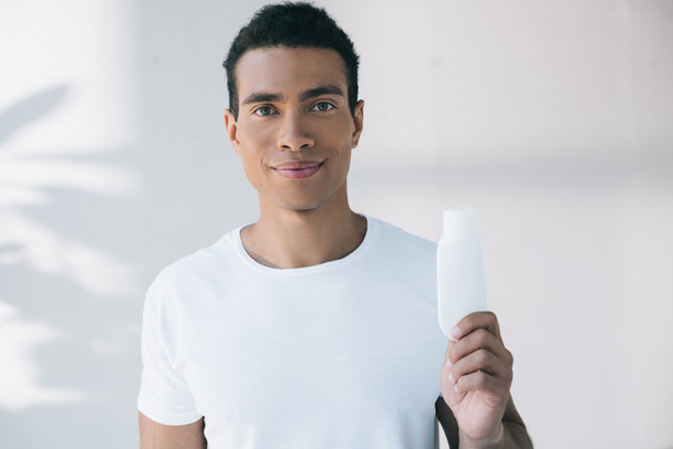 young man holding white bottle with cosmetic cream and looking at camera - Foto, imagen