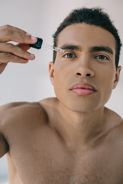 portrait shot of mixed race man dropping some serum on face with pipette while looking at camera - Фото, изображение
