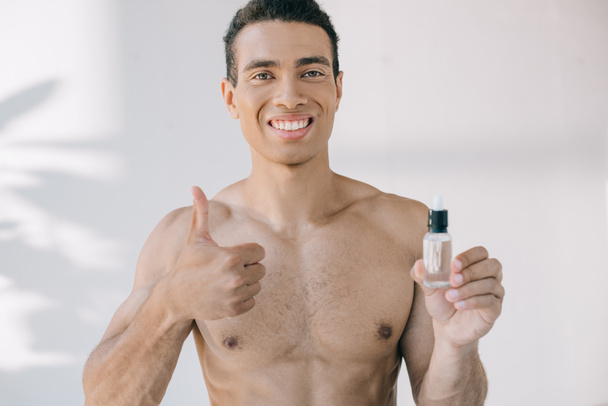 mixed race man holding bottle with serum and showing thumb up while looking at camera  - Fotó, kép
