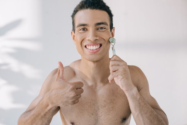 handsome mixed race man massaging face with jade roller and showing thumb up while looking at camera - Fotografie, Obrázek