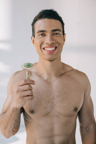 handsome muscular mixed race man presenting stone jade roller and smiling while looking at camera - Fotografie, Obrázek
