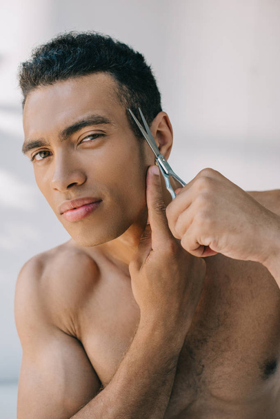 serious mixed race young man cutting temple with scissors and looking at camera - Φωτογραφία, εικόνα