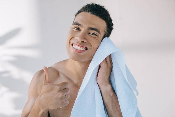 gespierde gemengde race man vegen gezicht met blauwe handdoek, weergeven van duim omhoog, glimlachen en kijken naar camera - Foto, afbeelding