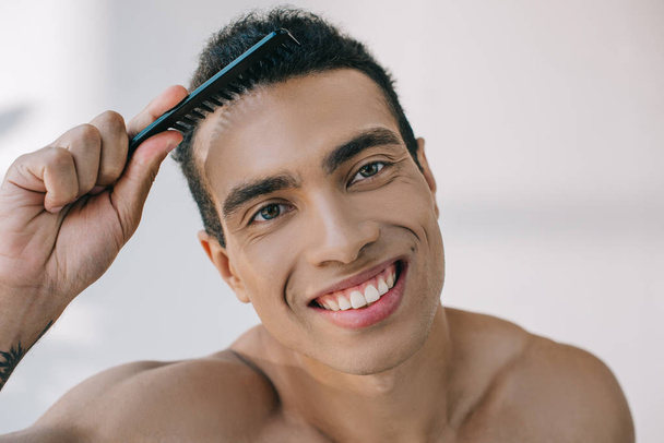 portrait shot of mixed race man combing hair with hairbrush and smiling while looking at camera - Foto, Imagen