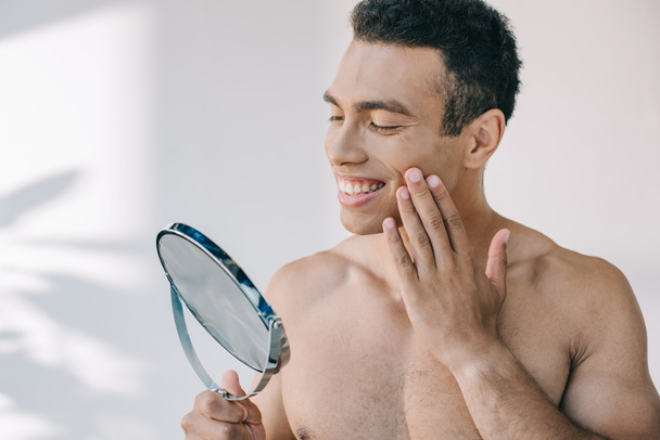 handsome mixed race man touching face with hand while looking at mirror - Foto, afbeelding