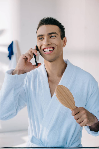 csinos férfi fürdőköpeny gazdaságban hajkefe és beszélgetett okostelefon, miközben mosolyogva és néz el - Fotó, kép