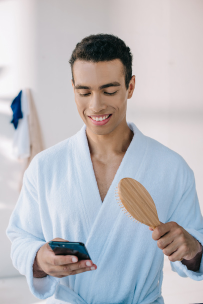 bell'uomo in accappatoio che tiene spazzola per capelli e smartphone mentre sorride e distoglie lo sguardo
 - Foto, immagini