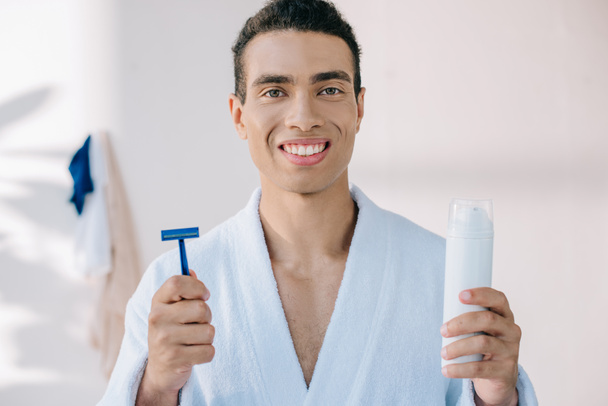 handsome young man in bathrobe holding razor and dispenser with shaving cream while looking at camera - Фото, изображение