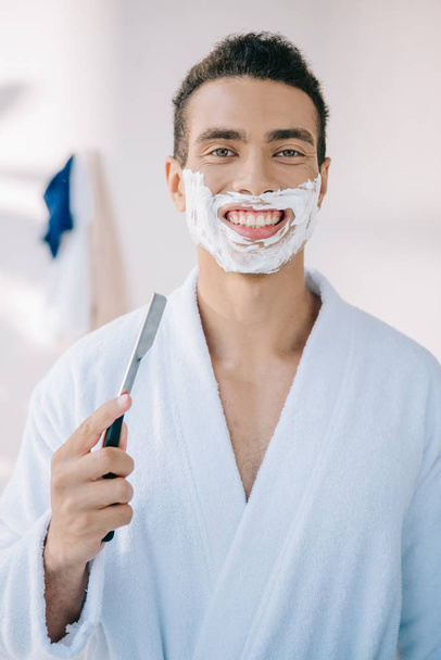 joven guapo con crema de afeitar en la cara sosteniendo hoja de afeitar, sonriendo y mirando a la cámara
 - Foto, imagen