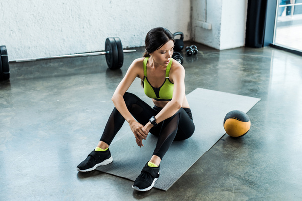 attractive sportswoman sitting on fitness mat and looking at ball  - Photo, Image