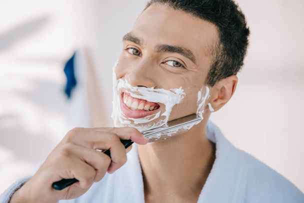 handsome young man in bathrobe shaving face with razor blade, smiling and looking at camera - Fotografie, Obrázek