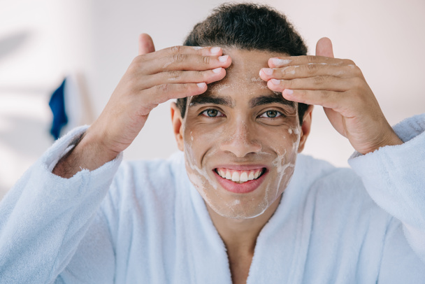 schöner Mann im Bademantel wäscht Gesicht mit Gesichtsschaum und lächelt in die Kamera - Foto, Bild