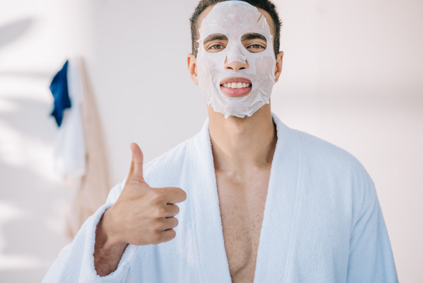 young man in bathrobe with face mask showing thumb up and looking at camera - Photo, Image