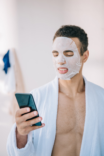 jovem em roupão de banho com máscara cosmética no rosto segurando smartphone
  - Foto, Imagem