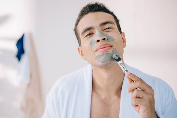 young man in bathrobe applying grey face mask with brush - Foto, Imagem