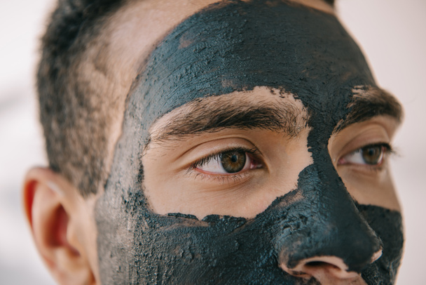 close up view of handsome young man with grey clay mask on face looking away - Foto, Imagen